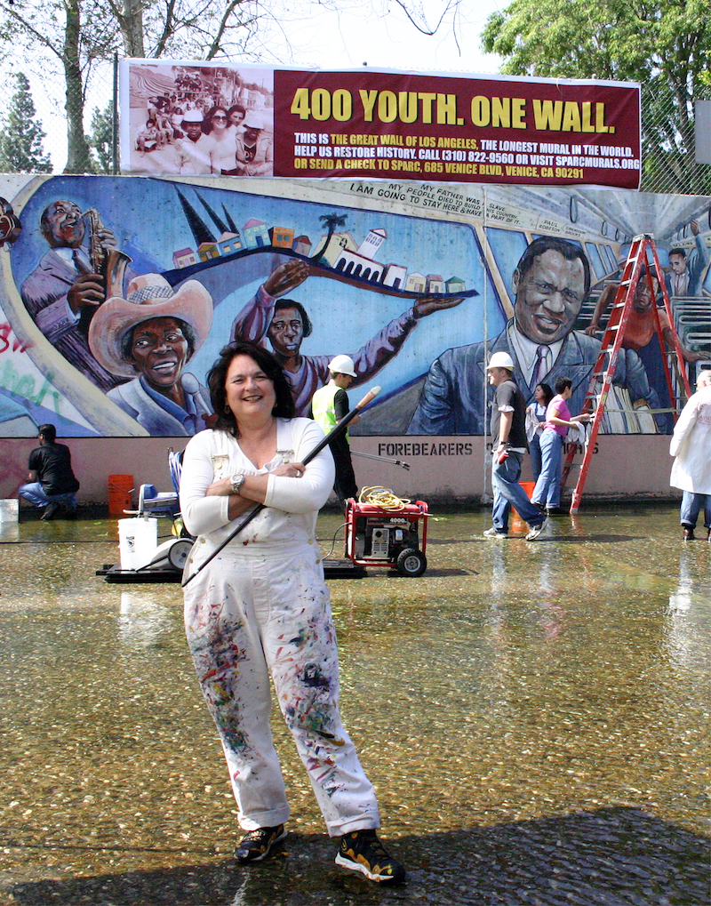 Judy Baca in a white jumpsuit in front of the Great Wall of Los Angeles