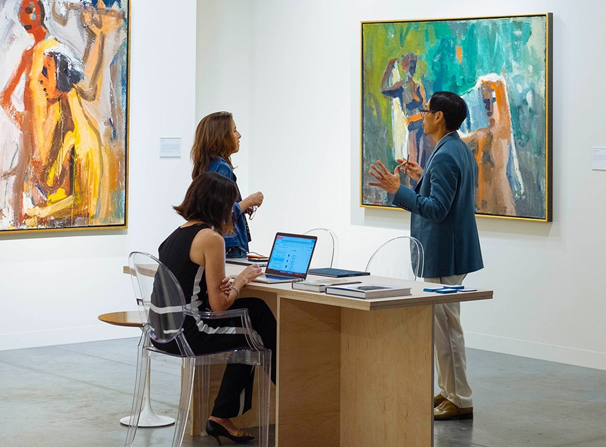 A woman is seated at a desk while on her laptop. A man and a woman are having an animated conversation beside her. Two large paintings are on the walls behind them.