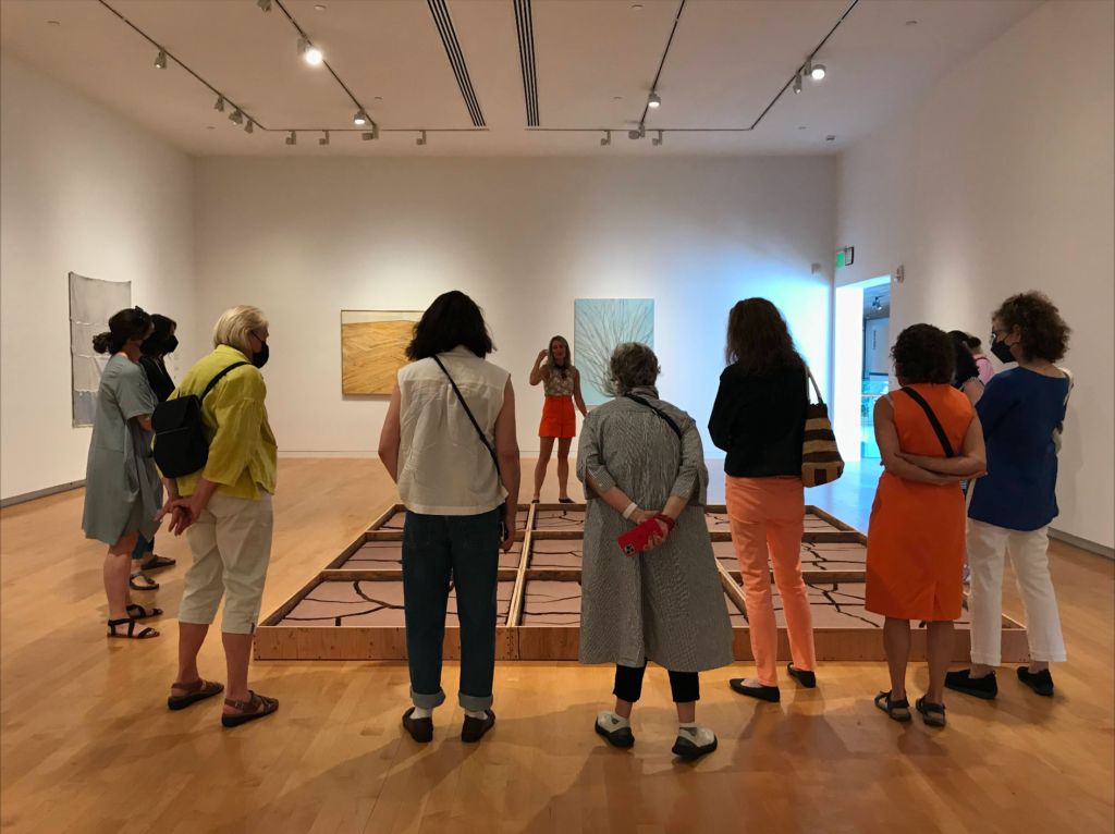 About 8 women are photographed from behind as they view a large artwork that is placed on the floor in front of them. The tour guide stands facing us on the other end of the artwork. The group is in a large room with white walls and some other paintings can be seen on the surrounding walls.