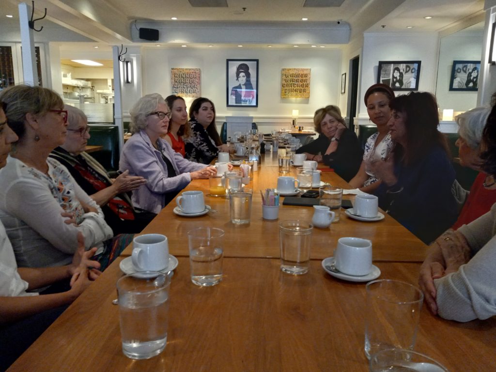 Women sit around a long table listening to one woman who is speaking. Coffee cups and glasses are on the table.